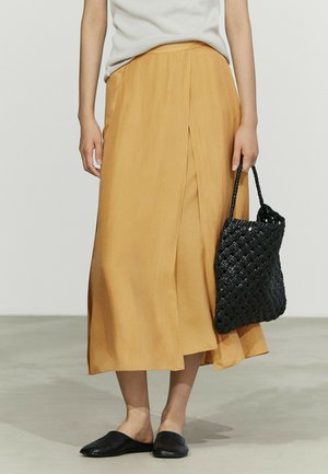 Donna che indossa una gonna midi giallo senape, top bianco senza maniche, scarpe slip-on nere, che tiene una borsa intrecciata nera al suo fianco.