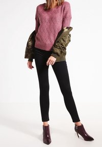 Maglione ribbed in mauve con un motivo testurizzato, abbinato a jeans skinny neri e stivaletti borgogna. Giacca bomber verde oliva poggiata sulla spalla.