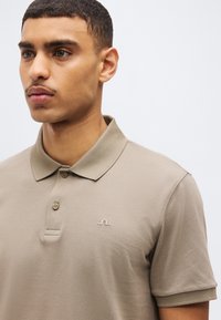 Νεαρός άνδρας με κοντά μαλλιά, που φοράει ένα μπεζ κοντομάνικο πόλο πουκάμισο με δύο κουμπιά και ένα μικρό λογότυπο στο στήθος.