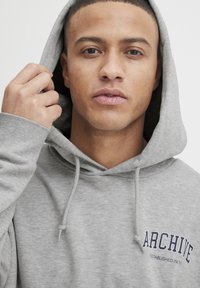 Grauer Sweatshirt mit Kapuze, mit einem Frontlogo in Marineblau, das "ARCHIVE ESTABLISHED IN 19XX" liest, weicher Textur und Kordelzügen.