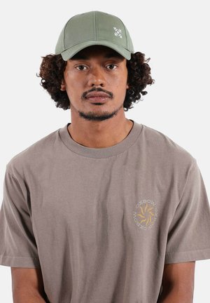 Homme aux cheveux bouclés portant une casquette vert clair et un t-shirt gris avec un petit logo circulaire jaune et blanc sur la poitrine.