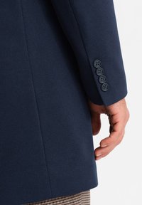 Giacca tessutata blu navy con finitura liscia, dotata di un polsino a quattro bottoni e una vestibilità curata e pulita. La manica presenta un delicato dettaglio di cucitura.