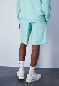Pantalones cortos de algodón color verde menta con un corte relajado y cintura elástica, combinados con zapatillas blancas y calcetines acanalados. Textura suave, sin patrones.