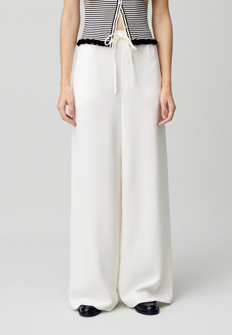 Pantalones anchos blancos con una textura suave y cintura con cordón. Combinados con una camiseta ajustada de rayas blancas y negras con un dobladillo con volantes.