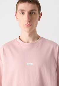 Light pink T-shirt made of cotton. Features a crew neck and a small white rectangular logo with "HUGO" on the front. Smooth texture.