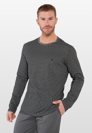 Man draagt een zwart-wit patroon lange mouw shirt en grijze broek, met één hand in de zak, staand tegen een effen lichte achtergrond.