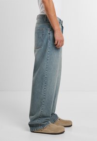 Világoskék denim, széles szárú farmernadrág, elhasználódott hatással, öt zsebes kialakítással és látható varrási részletekkel, bézs lábbelivel párosítva.