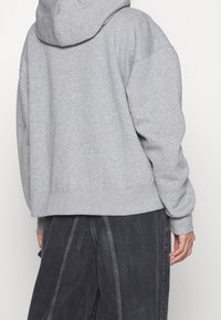 Vista trasera de una persona que lleva puesta una sudadera con capucha gris claro y pantalones oscuros gris sueltos con costuras visibles.