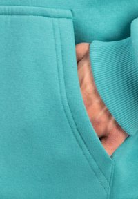 Felpa turchese con tasca a marsupio; tessuto in cotone texturizzato e polsini a coste; mano visibile all'interno della tasca.
