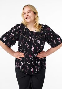 Blusa nera a fiori con maniche corte a palloncino, caratterizzata da un motivo floreale nei colori pastello e dettagli in pizzo lungo il collo.