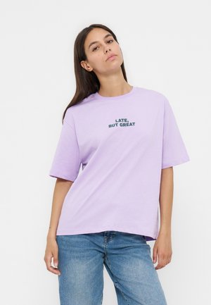 Jeune femme portant un t-shirt violet clair avec le texte « EN RETARD, MAIS GÉNIAL », associé à un jean bleu, debout devant un fond blanc.