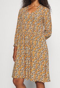 Blomstret kjole med lange ærmer, V-halsudskæring og indsnævret talje. Har et mønster af orange blomster på en mørk baggrund. Letvægtsstof.