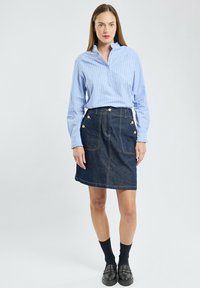 Chemise à rayures bleues avec col boutonné, associée à une jupe en denim foncé avec des accents dorés et des poches devant. Mocassins noirs et chaussettes.