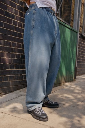 Person wearing baggy light blue jeans and black hiking shoes with gray laces standing on a sidewalk near a brick wall.