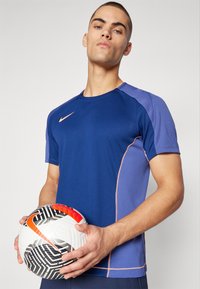 Camiseta atlética de manga corta en azul marino y morado con textura de malla y detalles en naranja, sostenida con un balón de fútbol de patrón en blanco y negro.