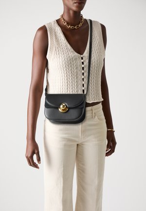 Borsa a tracolla nera in pelle con flap curvo, dettagli in hardware dorato e una lunga tracolla regolabile. Indossata su un maglione lavorato a maglia color crema.