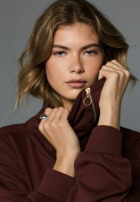 Tamsiai raudonas džemperis su aukštu apykakliu, užtrauktuko detale ir ribotais rankogaliais. Modelis laiko apykaklę, pabrėždamas švelnią tekstūrą ir minimalistinį dizainą.