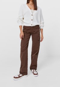 Bílý pletený kardigan s oversized rukávy a třemi knoflíky, kombinovaný s hnědými cargo kalhotami s bočními kapsami a bílými-hnědými teniskami.