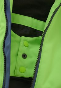 Inre i limegrön jacka med herringbone-textur, med en svart dragkedja och tryckknappsstängning, samt kontrasterande blå kant.