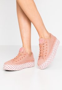 Różowe siateczkowe sneakersy na białej teksturowanej platformie z czerwonymi geometrycznymi wzorami, okrągłymi sznurowadłami i oznaczeniem marki na języku.