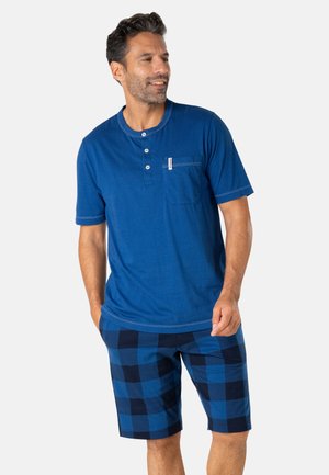 Homme portant un t-shirt Henley bleu à manches courtes avec une poche poitrine et un short à carreaux bleu et noir, debout les mains dans les poches.