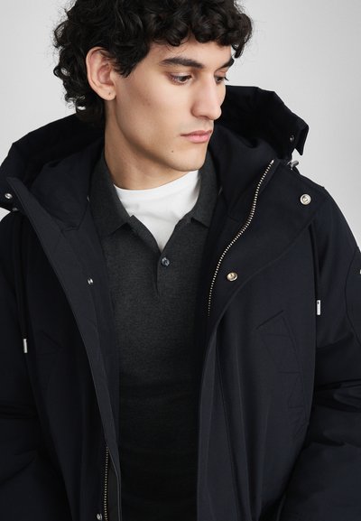 Jūrų laivyno parka su užtrauktuku priekyje, gobtuvu ir sidabrinėmis detalėmis, uždėta ant tamsiai pilko polo marškinėlių su tekstūriniais raštais.