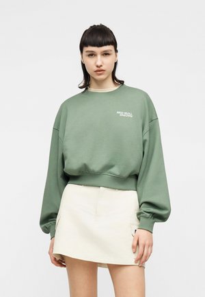 Ung kvinde med glat mørkt hår iført grøn Red Bull Racing-sweatshirt og hvid mininederdel, stående mod en ensfarvet baggrund.