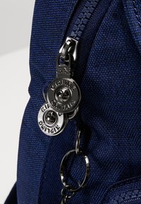 Sac en tissu bleu marine à surface texturée, doté d'une fermeture éclair noire proéminente et de deux curseurs métalliques gravés du mot "Kipling".