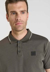 Camisa polo de color verde oliva oscuro con una textura sutil, cierre de tres botones y un parche con logo. Presenta detalles en blanco en el cuello.