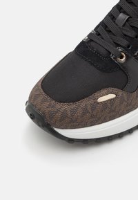 Primer plano de la puntera de una zapatilla negra y marrón con un ribete lateral estampado, cordones negros y suela blanca, sobre un fondo blanco liso.