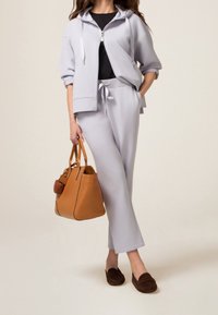 Světle šedá mikina se zipem vpředu, odpovídající teplákové kalhoty a hnědé mokasínové pantofle. Hnědá kabelka s kulatým akcentem.