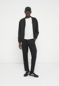 Μαύρο μπουφάν με φερμουάρ πάνω από γκρι φούτερ, συνδυασμένο με μαύρο παντελόνι και μαύρα αθλητικά παπούτσια με λευκές ρίγες. Το μοντέλο φοράει μαύρο καπέλο.