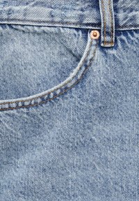Acercamiento al bolsillo de unos jeans de mezclilla azul claro con costuras naranjas y un remache de cobre cerca del lazo del cinturón.