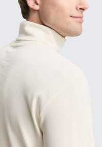 Mann in einem cremefarbenen Rollkragenpullover, der sich leicht dreht und die Seite seines Gesichts sowie den oberen Rücken zeigt.