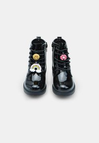 Botines de charol negros con cordones, que presentan coloridos charm de plástico: una cara sonriente, un arcoíris y una flor rosa en la parte frontal.