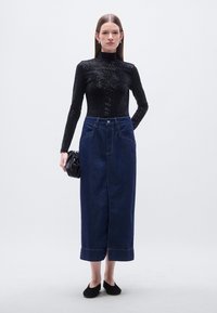 Μαύρη μπλούζα με μακριά μανίκια και υφή υφάσματος· σκουρόχρωμη φούστα από τζιν με ψηλή μέση, τσέπες και εμφανές ράψιμο· μοντέλο κρατά μικρή μαύρη τσάντα.