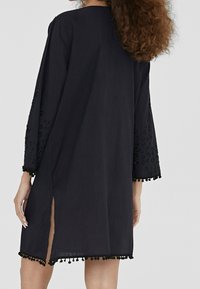 Svart knälång tunika med 3/4-ärmar. Har broderade detaljer och pom-pom kant på ärmarna och fållen. Lättviktsmaterial, sidofspring.