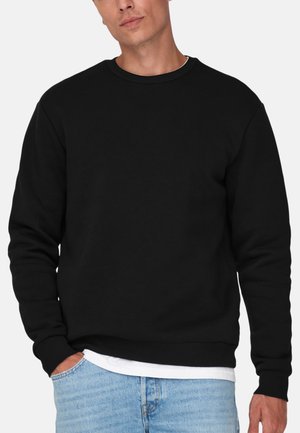 Mand iført en ensfarvet sort crewneck sweatshirt over en hvid skjorte og lyseblå jeans, stående med den ene hånd i lommen.