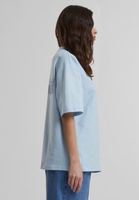 Camiseta de algodón azul claro con mangas cortas, corte relajado y detalle de texto en la parte posterior. Combinada con jeans de mezclilla azul.