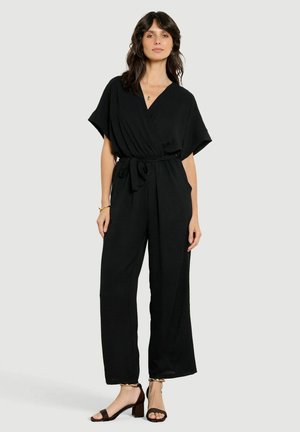 Vrouw met donker haar die een zwarte jumpsuit met riem en donkere open-toe hakken draagt, staand tegen een eenvoudige lichte achtergrond.