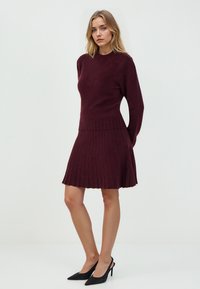 Burgunderfarbendes Strickkleid mit langen Ärmeln, gerippter Textur und schwingendem Rock; Saum mit Fransen-Detail. Kombiniert mit schwarzen spitzen Stiletto-Heels.