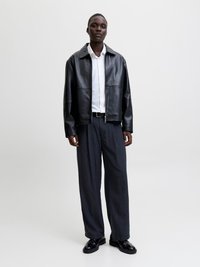 Άτομο που στέκεται φορώντας μαύρο δερμάτινο μπουφάν, λευκό πουκάμισο με κουμπιά, σκούρα παντελόνια με ρίγες και φαρδύ πόδι, και μαύρα επίσημα παπούτσια μπροστά σε απλό φόντο.