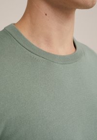 Světle zelený crewneck svetr, bavlněná látka, hladká textura, vroubkovaný límec, detailní záběr na oblast výstřihu a ramene, bez vzorů nebo akcentů.