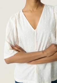 White, textured blouse featuring a V-neck, short puff sleeves, and subtle raised dot patterns. Lightweight fabric with a relaxed fit.