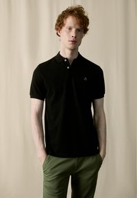 Camisa polo negra con cuello clásico, que presenta un pequeño logo bordado en el pecho. La tela parece suave y ligera.