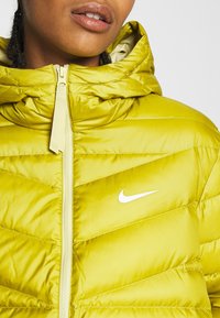 Limenvihreä puhvijakku, jossa on vaakasuora tikattu kuvio. Varustettu hupulla, täyspitkällä vetoketjulla ja valkoisella Nike-logolla rinnassa. Pehmeä tekstuuri.