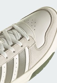 Gros plan d'une basket blanche avec des empiècements en suède, une boîte à orteils perforée, trois bandes grises et blanches, et des lacets blancs sur un fond clair.
