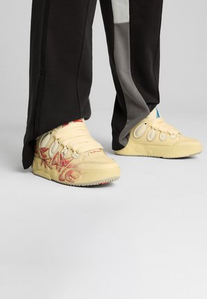 Gelbe Sneakers mit strukturierten Details, kreisförmigen Akzenten und roten Grafiken. Kombiniert mit schwarzen Jogginghosen mit seitlichen Akzenten.