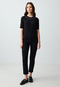 Zwarte, kortärmed t-shirt met een ronde halslijn, versierd met decoratieve parelaccenten. Gecombineerd met strakke zwarte broeken en zwarte instapschoenen.
