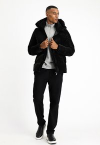Chaqueta acolchada negra con capucha, cierre de cremallera y tejido texturizado, llevada sobre una camiseta gris de cremallera, combinada con pantalones negros y zapatillas negras.